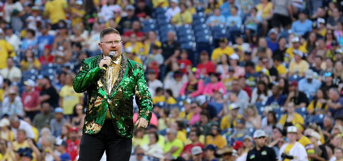 Rowan University alumnus Matt Graifer of the Savannah Bananas wears a green glittery suit jacket and holds a microphone with a stadium full of fans behind him.