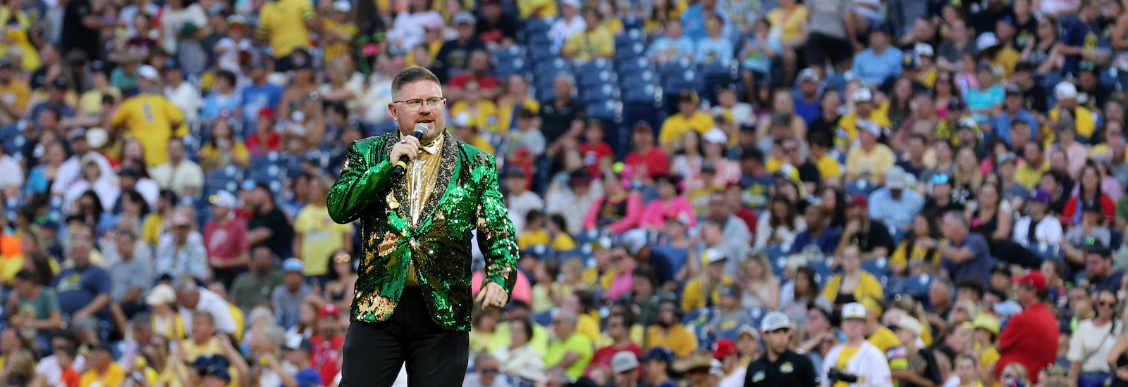 Rowan University alumnus Matt Graifer of the Savannah Bananas wears a green glittery suit jacket and holds a microphone with a stadium full of fans behind him.