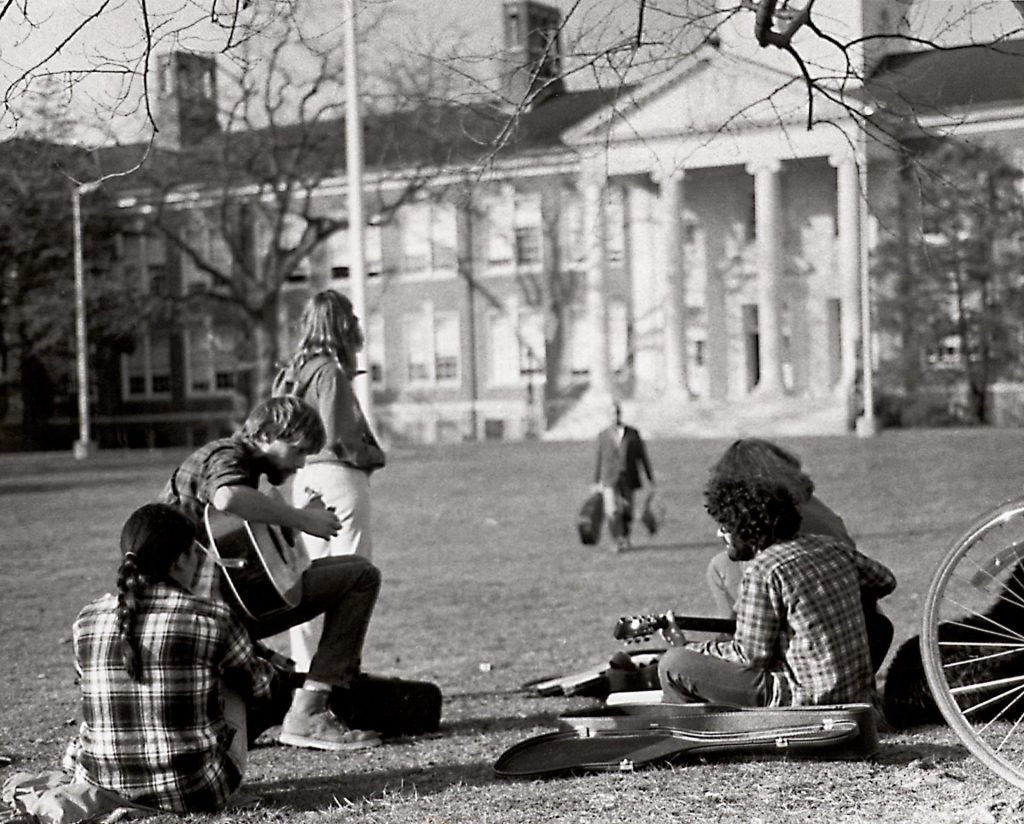 From the 1970s historical archives of Rowan University, a professor walks toward students sitting casually on Bunce Green. 