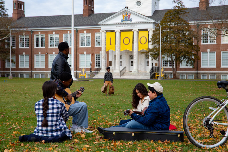 Six Rowan University students sit on Bunce Green with hats, bicycles, briefcases and guitars that perfectly recreate a black and white photo from the 1970s. 