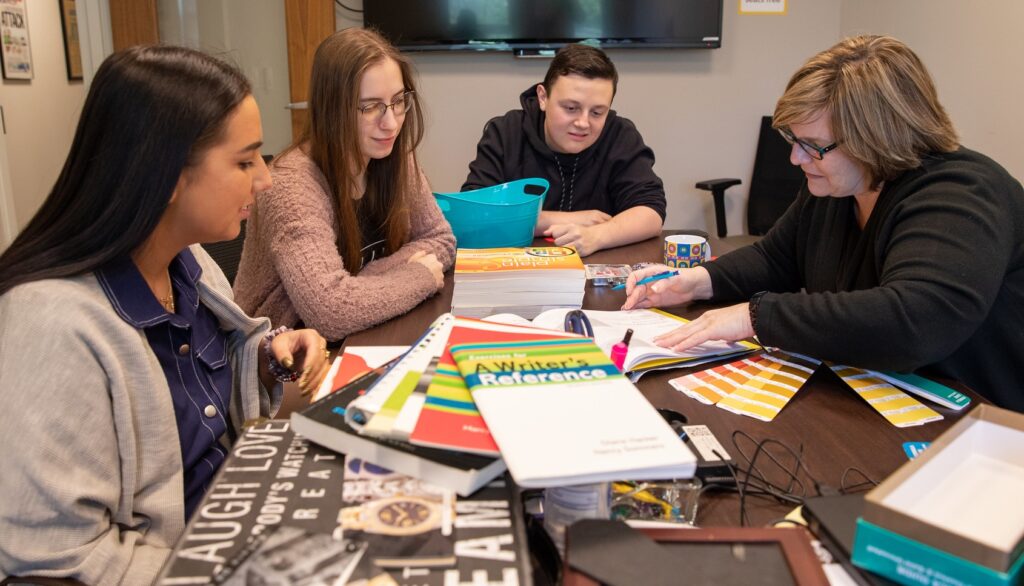 Rowan University advertising professor Lisa Fagan sits as a messy, full table, working hands-on with three students who are intently watching what she is pointing out. 