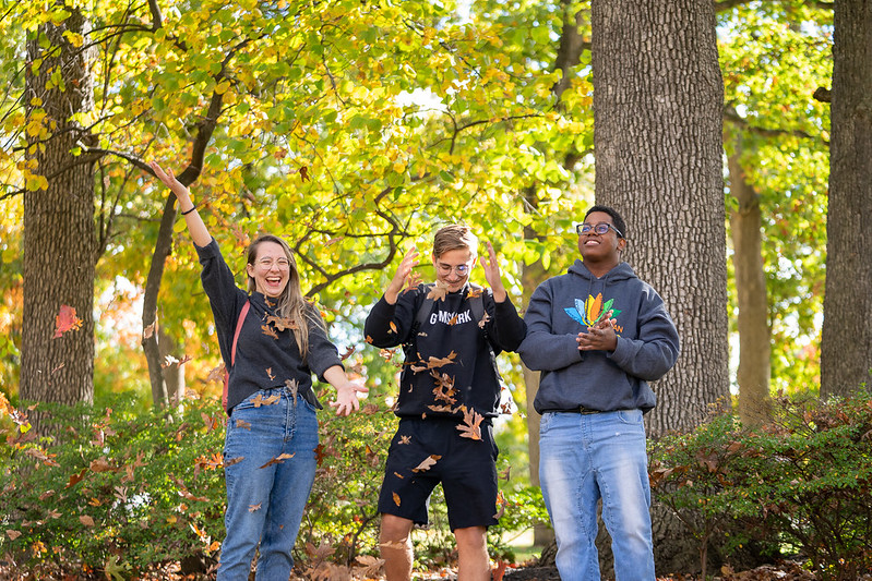 Three Rowan University students stand in a wooded area of campus, playing with autumn leaves by throwing them in the air. 