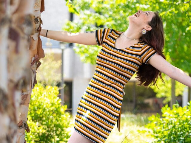 A Rowan University holds onto a tree, leaning backward with one arm hanging freely, laughing.