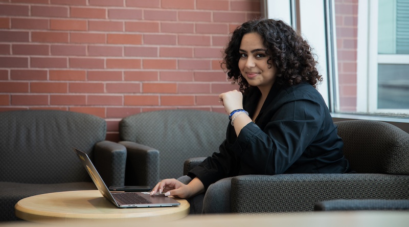 A Rowan University student dressed in a dark business casual outfit sits alone working on a laptop. 