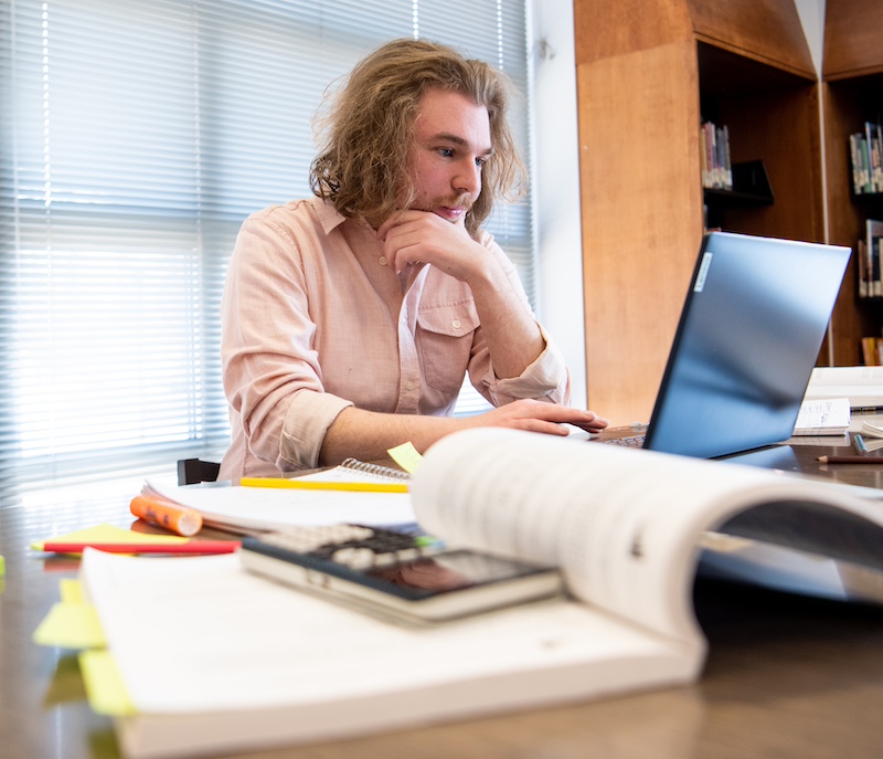 A Rowan University student works on his laptop, surrounded by books, sticky pads, a highlighter and his phone. 