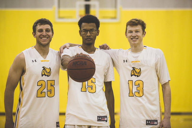 Three Rowan Men's basketball team members stand shoulder to shoulder, with the one in the center holding a basketball out straight forward.