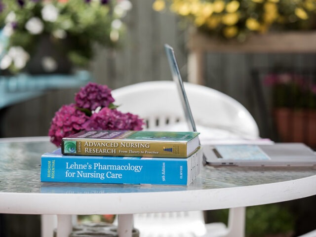 Two master of nursing textbooks sit on an outdoor patio table, with an open laptop and a bouquet of dark pink flowers.