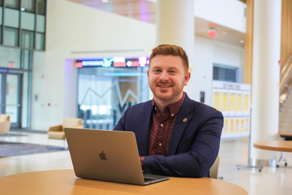Male business student seated at a table with an open laptop in a university building.