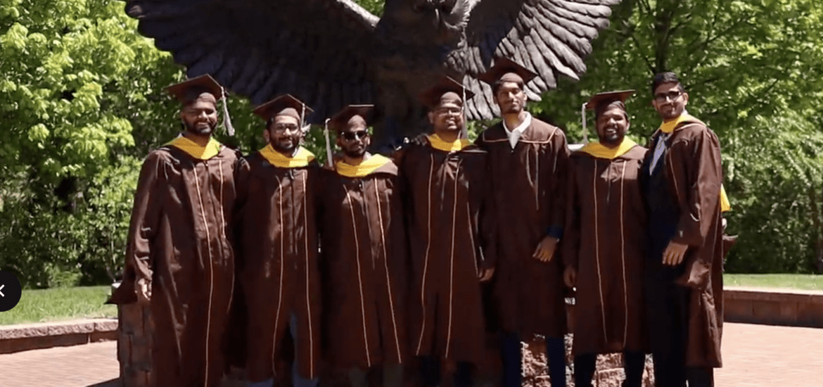 Indian friends at Rowan University stand in their graduation cap and gown attire in front of a statue of the school mascot.