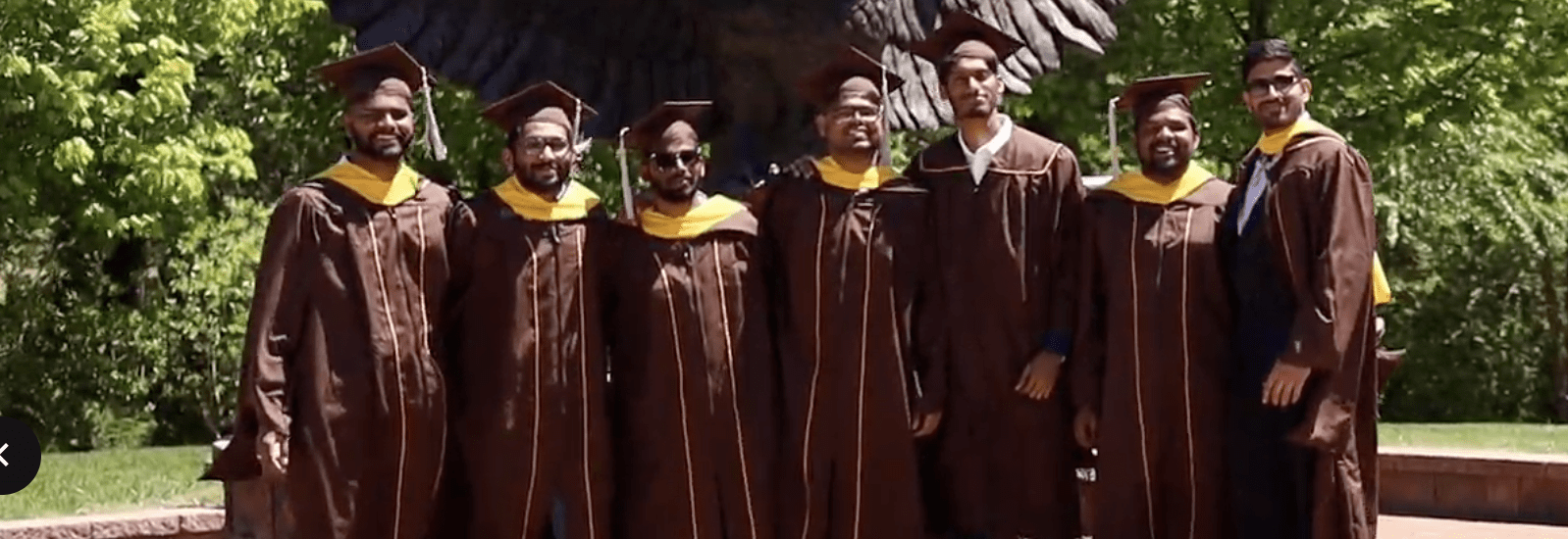 Indian friends at Rowan University stand in their graduation cap and gown attire in front of a statue of the school mascot.