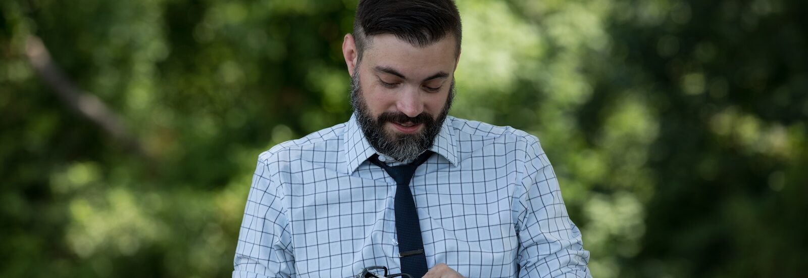 A Rowan University master of engineering management graduate stands outdoors while wearing a shirt and tie, cleaning his glasses.
