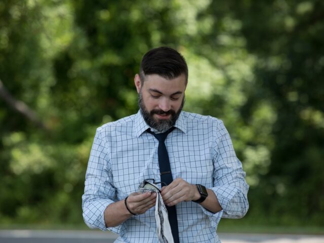 A Rowan University master of engineering management graduate stands outdoors while wearing a shirt and tie, cleaning his glasses.