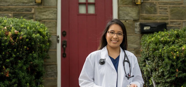 A Rowan University Rn to BSN online nursing student stands outside her home, with a red door behind her, wearing a white lab coat and with a stethoscope around her neck.