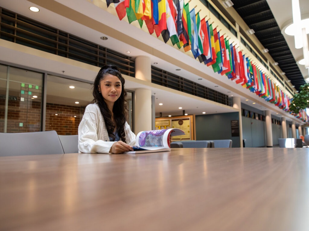 Rowan University student sitting inside James Hall at a table reading a magazine with a row of international flags behind her.