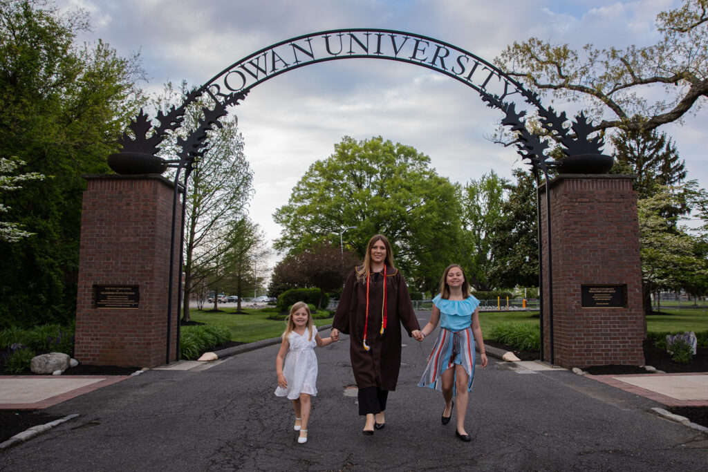 Rowan online graduate in cap and gown walking through campus arch with her two daughters