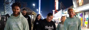 A group of young people walk together toward the photographer on a summer night boardwalk at the Jersey Shore, wearing Butterfly Effect apparel.
