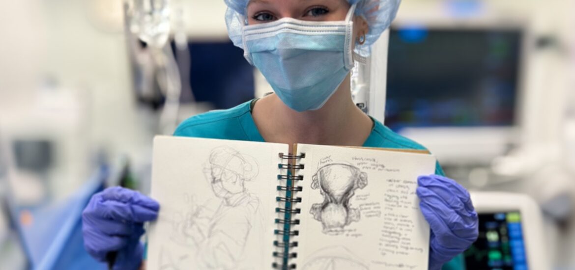 A Rowan University biomedical art and visualization major holds up a medical illustration in an operating room while wearing blue medical scrubs, gloves, mask and cap.
