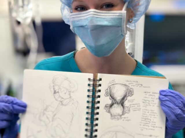 A Rowan University biomedical art and visualization major holds up a medical illustration in an operating room while wearing blue medical scrubs, gloves, mask and cap.