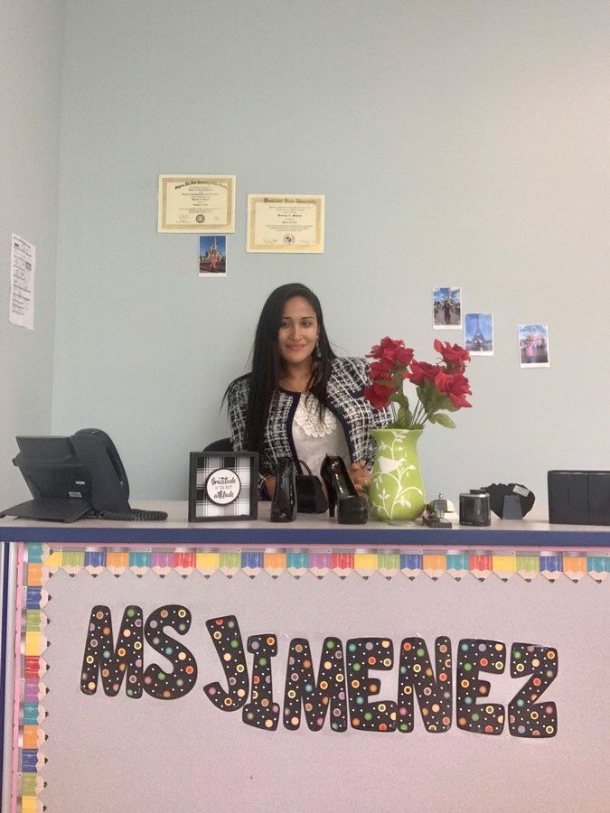 Manuela Jiménez, a Rowan Ed.D. student and full-time teacher, smiling behind her classroom desk labeled "Ms. Jimenez," decorated with flowers and classroom supplies