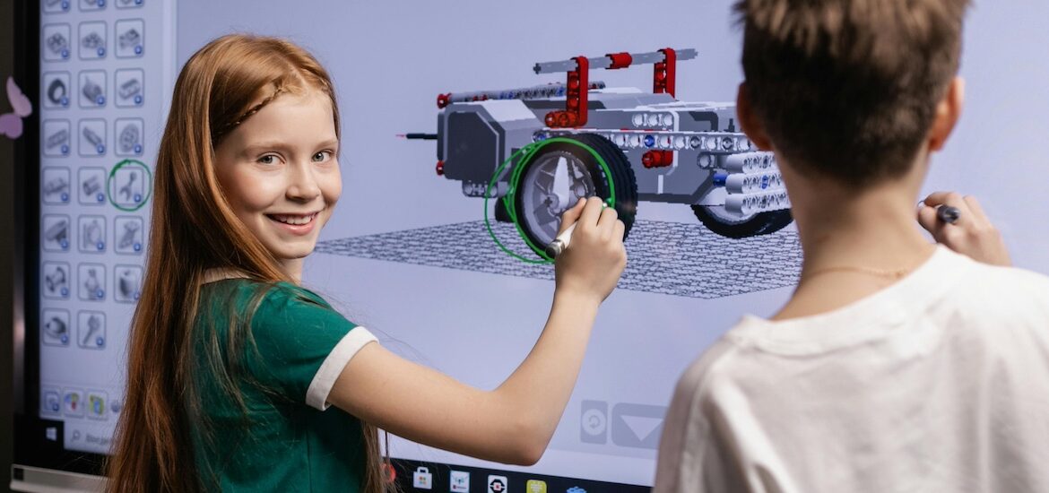 A middle school student looks over her shoulder at the camera smiling as she draws on a large interactive screen with another student.