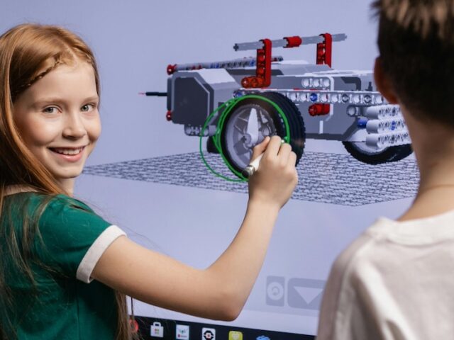 A middle school student looks over her shoulder at the camera smiling as she draws on a large interactive screen with another student.