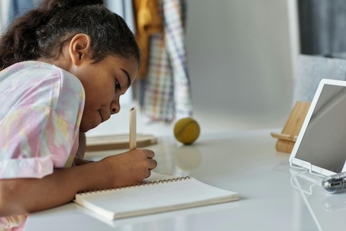 A young student leans forward on her desk to write in a notepad, with a tablet visible in front of her.