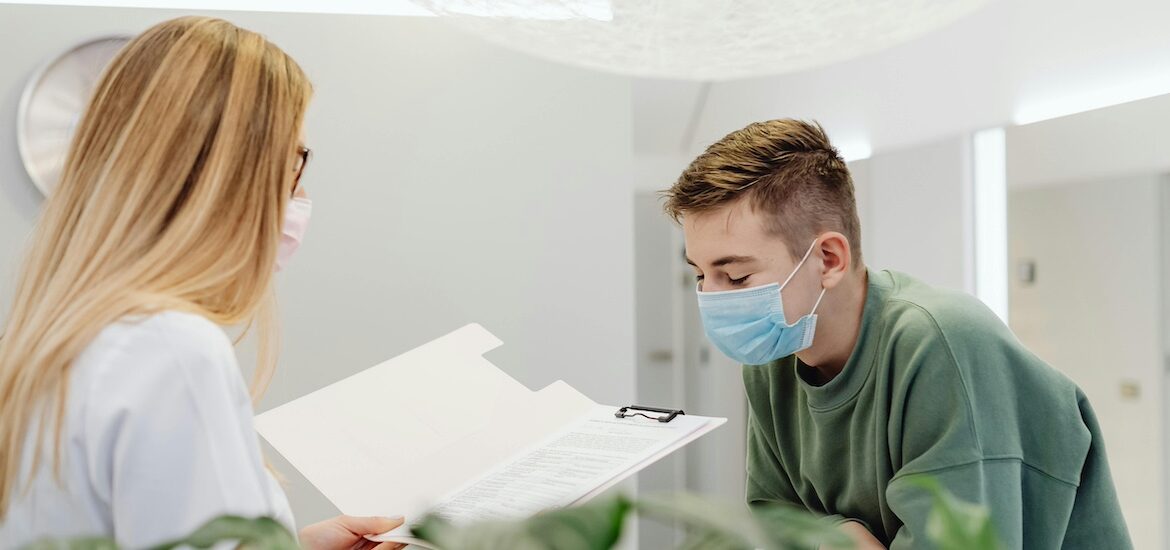 A healthcare administrator stands behind a desk reading something in a folder with a patient in front of her.
