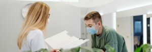 A healthcare administrator stands behind a desk reading something in a folder with a patient in front of her.