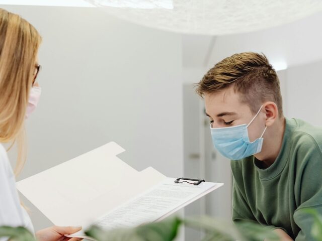 A healthcare administrator stands behind a desk reading something in a folder with a patient in front of her.