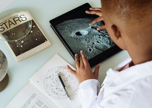 A middle school student studies craters using a book, a notepad and a tablet. He is zooming in on the tablet.