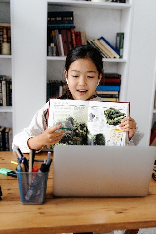 An elementary school student excitedly shows the inside of a book in view of her laptop to the person on the other end.
