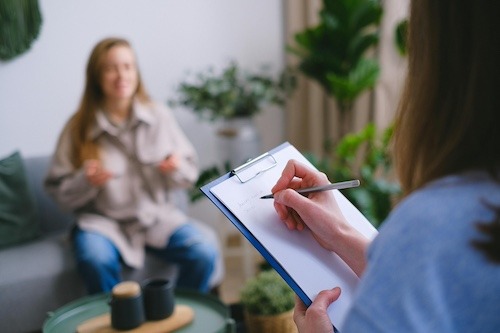 A healthcare worker writes notes on on a clipboard as the patient sits on a couch across from her.