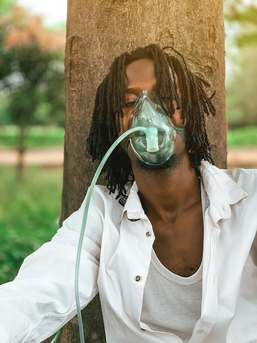 A man uses an oxygen mask while outside, sitting against a tree with his eyes closed.