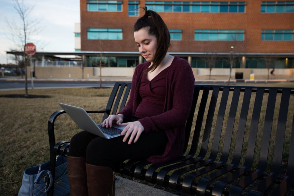 A Rowan University bachelor's of science in nursing BSN student sits on a bench in a park, looking down at her laptop perched on her lap.