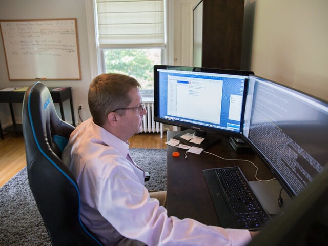 A cybersecurity professional works at a desk with two large computer monitors displaying technical information.