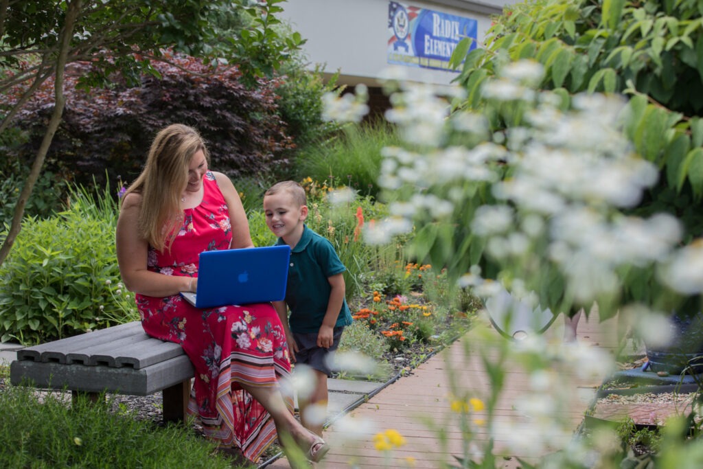 Woman completing online coursework on a laptop while spending time with her young son in a garden outside an elementary school