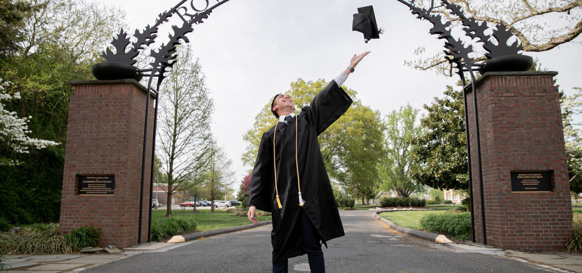Graduating Rowan University online student standing confidently under the Rowan arch.