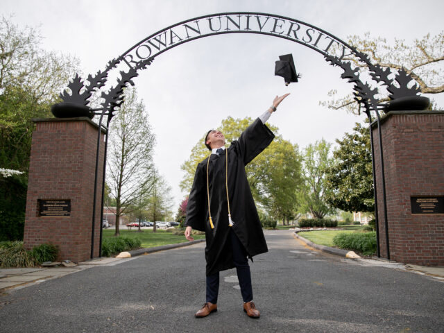 Graduating Rowan University online student standing confidently under the Rowan arch.