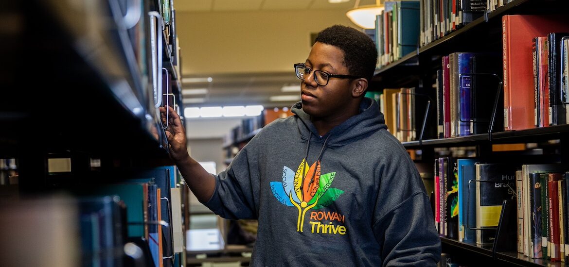 A Rowan University student browses books in the library wearing a Rowan Thrive shirt indicating the pillars of wellness at the university.