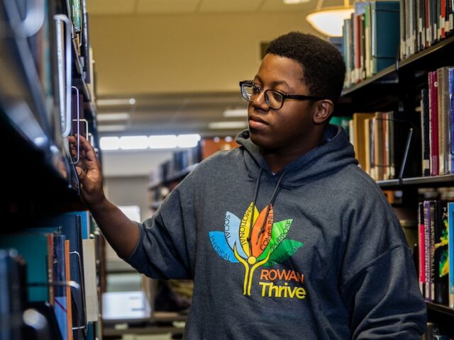 A Rowan University student browses books in the library wearing a Rowan Thrive shirt indicating the pillars of wellness at the university.