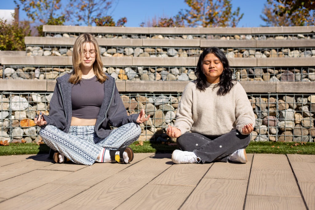 Two Rowan University students sit side by side outdoors in a seated meditation pose with eyes closed.