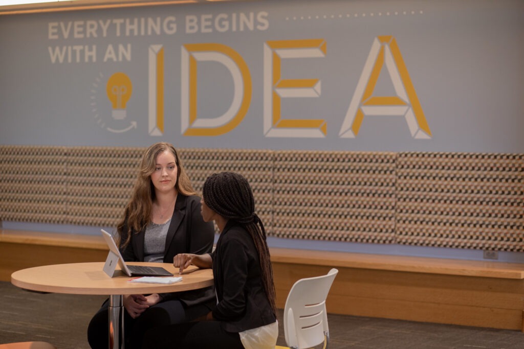 Two students discussing what data analytics is at a table with a laptop and an 'Everything Begins With an Idea' wall display