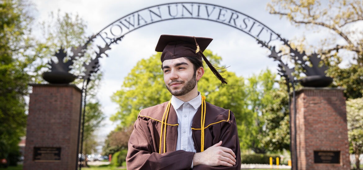 Graduating Rowan University online student standing confidently under the Rowan arch