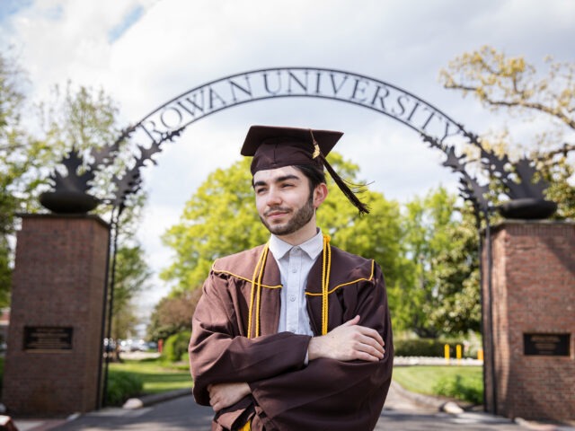 Graduating Rowan University online student standing confidently under the Rowan arch