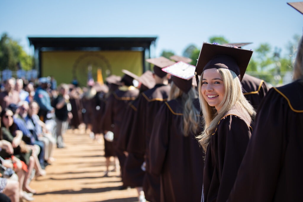 A smiling student in a graduation cap and gown looks back while walking with classmates at a commencement ceremony