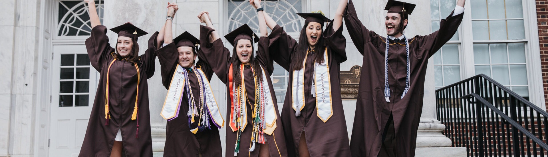 Group of college graduates in caps and gowns holding hands and cheering on graduation day, celebrating the excitement of commencement and new beginnings.