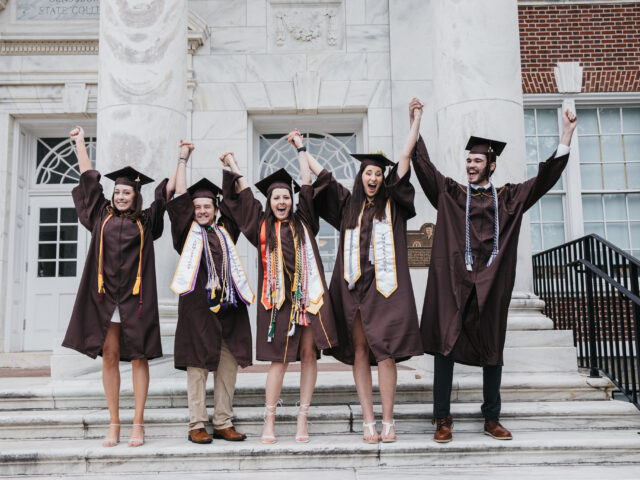 Group of college graduates in caps and gowns holding hands and cheering on graduation day, celebrating the excitement of commencement and new beginnings.