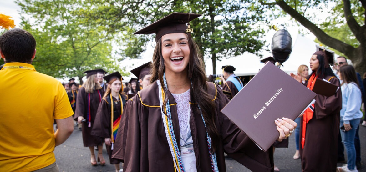 A smiling Rowan University graduate holds her diploma during commencement, representing pride, accomplishment, and the start of a new chapter