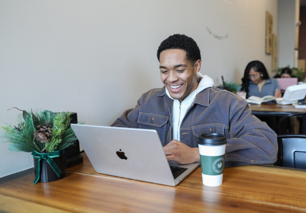 Recent graduate smiling while working on a laptop in a café, reflecting optimism and curiosity while planning next steps after college