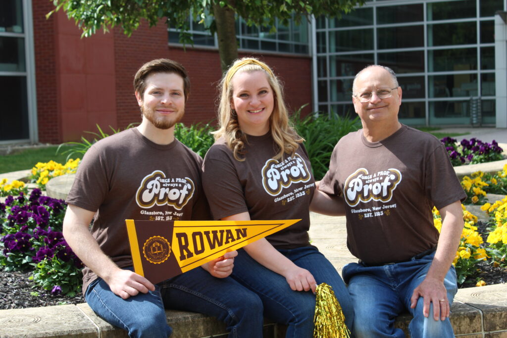 Three Rowan University alumni wear “Once a Prof, Always a Prof” shirts and smile while holding a Rowan pennant on campus